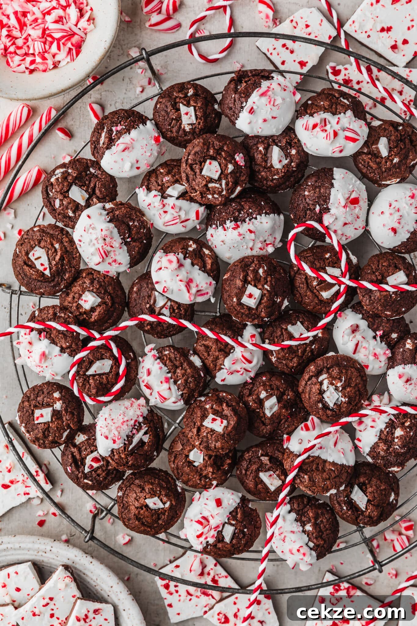 A wire rack of mini chocolate peppermint bark cookies on a grey counter next to a white bowl of chopped candy canes, a white bowl of peppermint bark, and striped red and white ribbon.