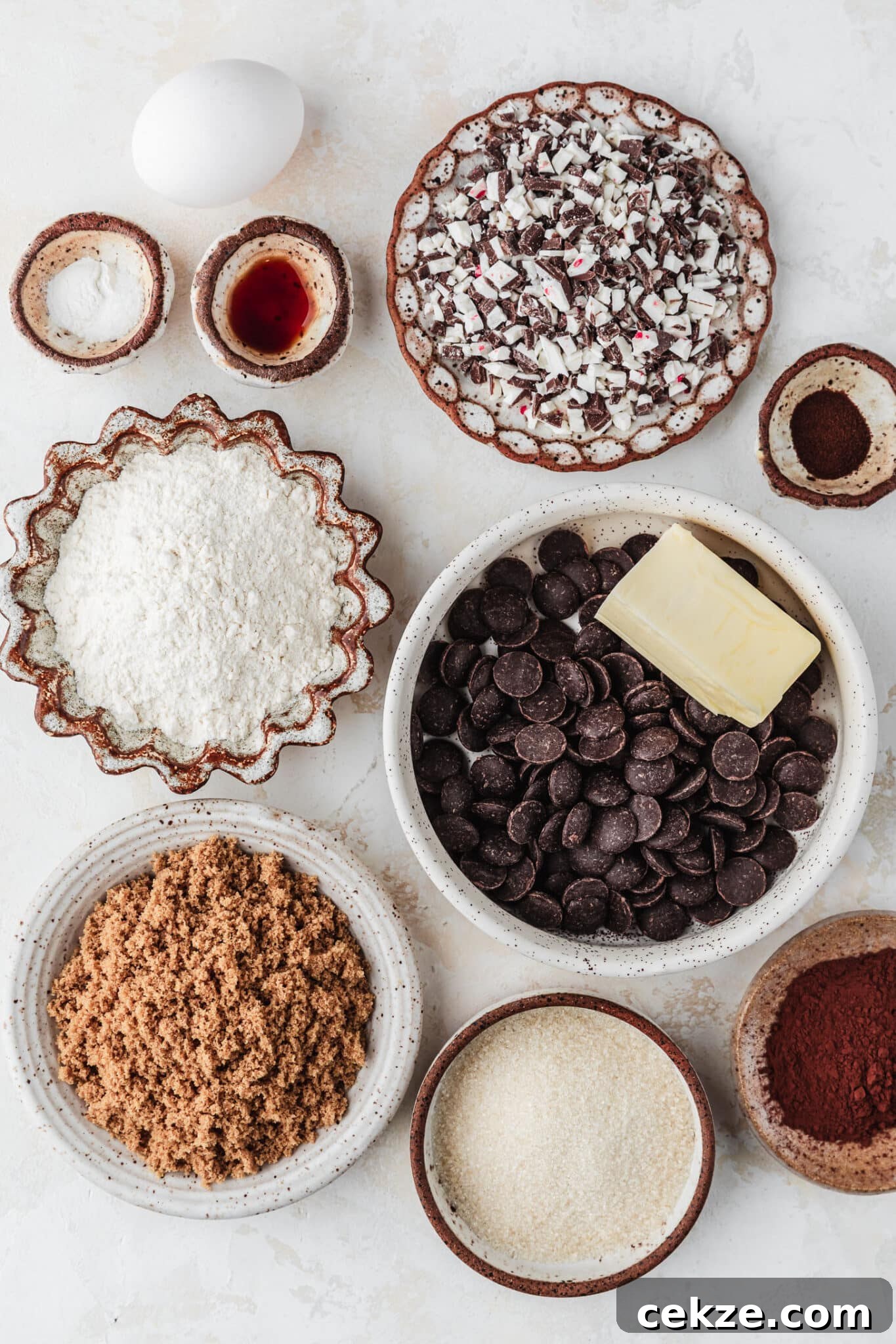 White and brown bowls of chocolate chips, butter, flour, brown sugar, white sugar, cocoa powder, vanilla, baking soda, espresso powder, and peppermint candy next to an egg on a tan counter.