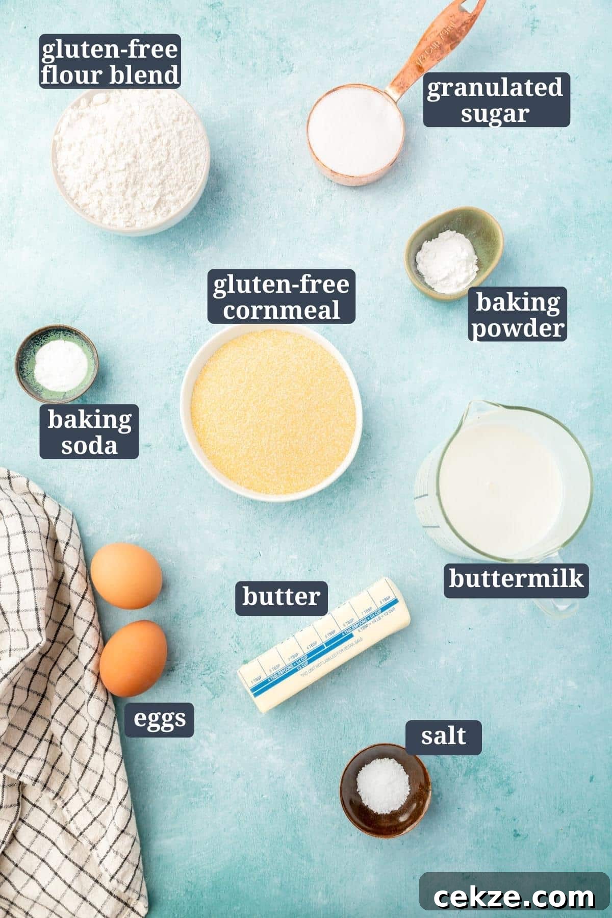 An overhead view of ingredients in small bowls on a blue table for making gluten-free cornbread, including gluten-free flour, sugar, salt, baking soda, baking powder, buttermilk, cornmeal, eggs, butter with text overlays over each ingredient.