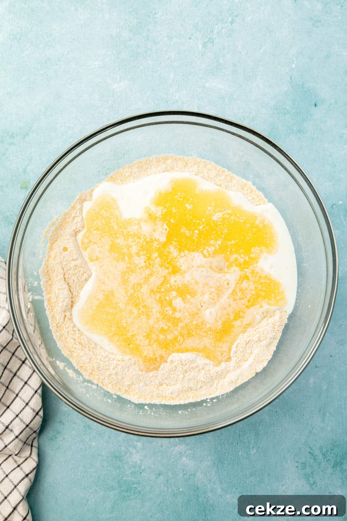 A glass mixing bowl with dry ingredients for cornmeal topped with buttermilk, whisked eggs, and melted butter.