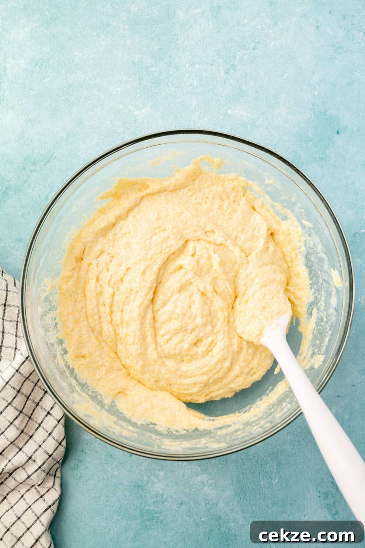 A glass mixing bowl with cornbread batter that is being mixed up with a spatula.
