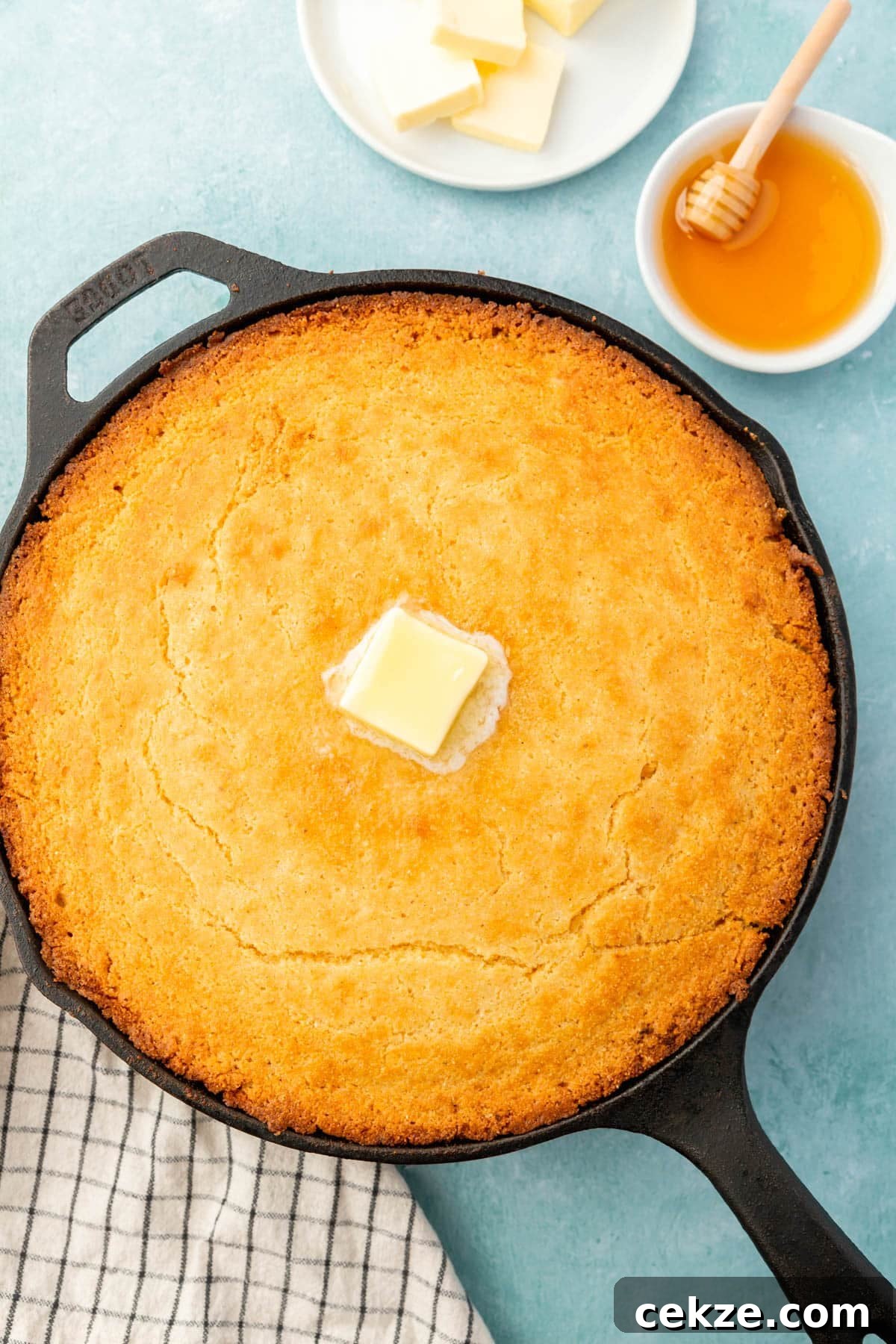 A cast iron skillet filled with gluten-free cornbread topped with a pat of melted butter with a ramekin of honey and a plate of butter on the side.