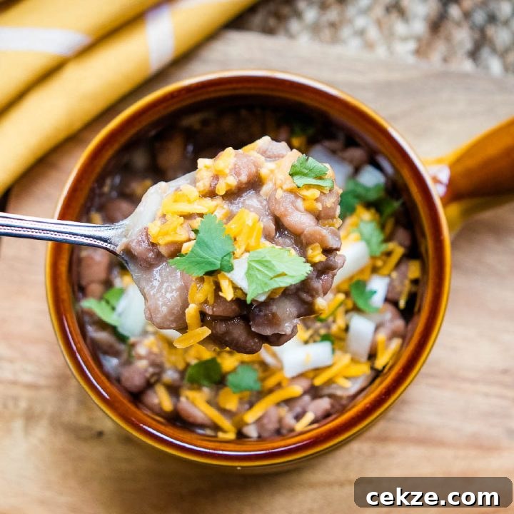 A spoonful of cooked pinto beans topped with shredded cheddar cheese and cilantro. Below the spoon is a bowl with a handle on a wooden cutting board and there is a yellow and white napkin on the edge of the cutting board. The bowl is full of cooked pinto beans topped with diced onions, shredded cheddar cheese, and cilantro.