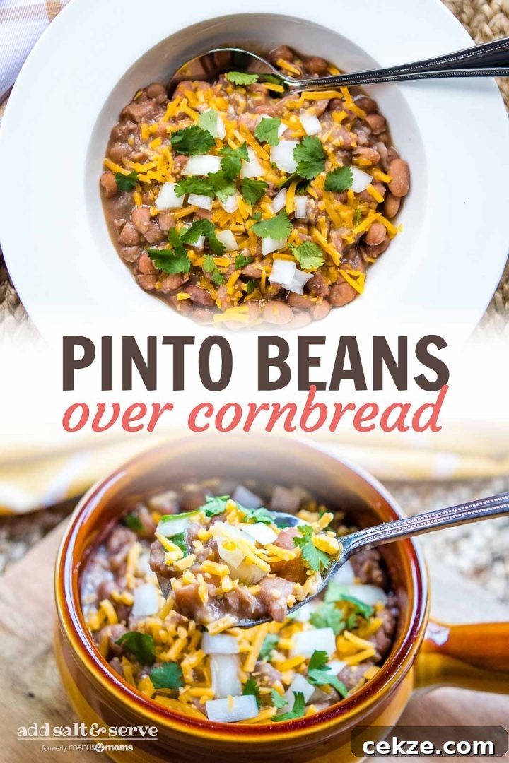 A spoonful of cooked pinto beans topped with shredded cheddar cheese and cilantro. Below the spoon is a bowl with a handle on a wooden cutting board and there is a yellow and white napkin on the edge of the cutting board. The bowl is full of cooked pinto beans topped with diced onions, shredded cheddar cheese, and cilantro.